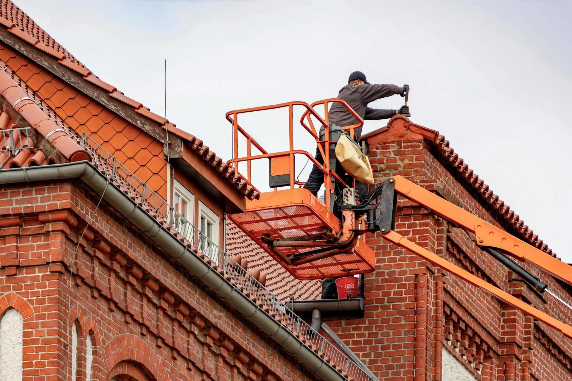 Un couvreur, à bord d'une nacelle élévatrice, effectue des travaux d'entretien sur un bâtiment en briques rouges au toit de tuiles.